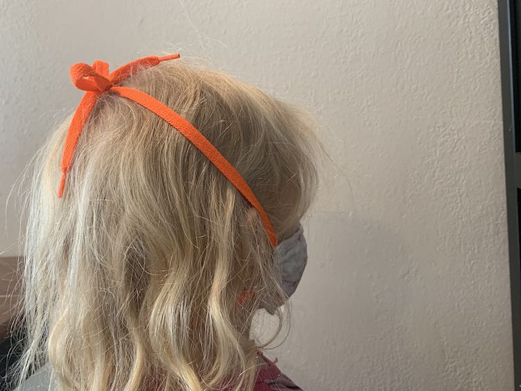 View from the side as the mask is worn and the lower loop goes behind Zoey's neck, under her hair, and the upper shoelaces extend over her ears and tie at the top of her head.