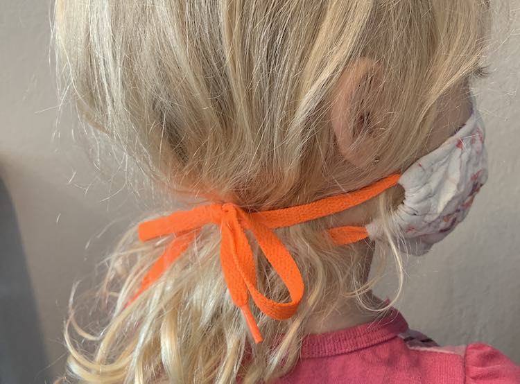Image shows the side of Zoey's face while she wears the face mask with the upper loops pulled under her ears and tied in the back at the nap of her neck over her hair. The lower loop is under her hair but both are looped at the same spot. 