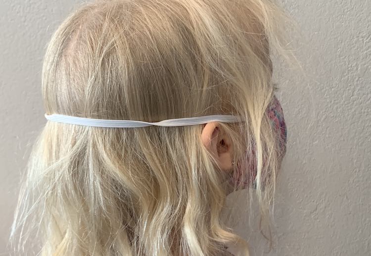 View of the back and side of Zoey's head while she's wearing a mask with elastic going around her head. The lower elastic is hidden under her hair but the top elastic band goes over her ears and around the back of her head. 