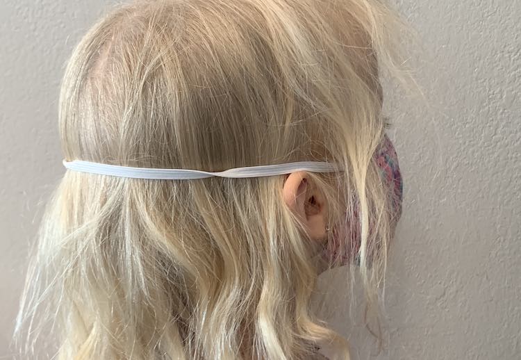 View of the back and side of Zoey's head while she's wearing a mask with elastic going around her head. The lower elastic is hidden under her hair but the top elastic band goes over her ears and around the back of her head.
