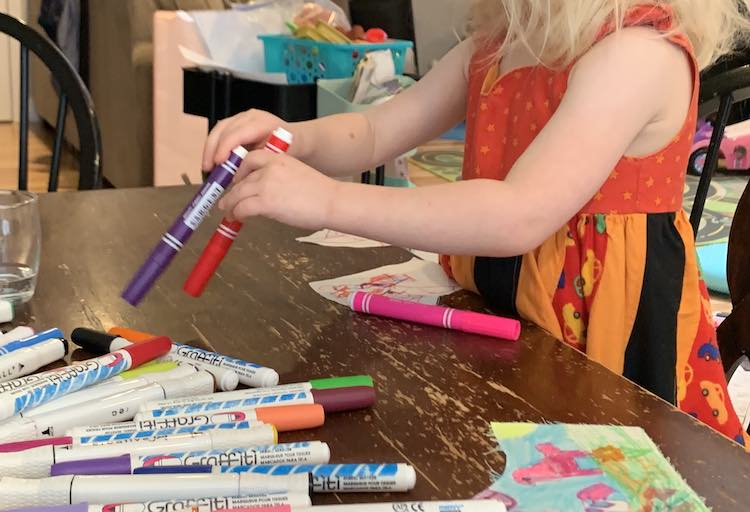 Image shows Zoey on her knees on a chair at the table. Her two mask pieces are in front of her as she reaches over them to grab two fabric markers from the pile in the center of the table. You can see part of one of Ada's pieces at the lower right side of the image.