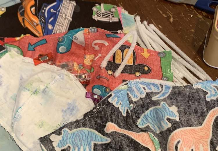 Image shows a pile of inside out face masks and white pipe cleaners. The back masks are right side out and slightly blurred. The center one is inside out with a seam allowance. The front left one has a pipe cleaner clipped to a mask while the mask on the right is sewn to the mask at the center and either end of the pipe cleaner. There's a pile of straight pipe cleaners to the side a one in the center folded in half so it could match the center with the center seam of the mask.
