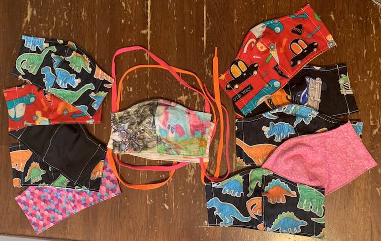 Image shows a colored mask in the center, with the other colored one hidden below. There are neon orange and pink shoelaces coming out of them from the side channels. To the left and right are ten other masks without any shoelaces or elastic in the channels.