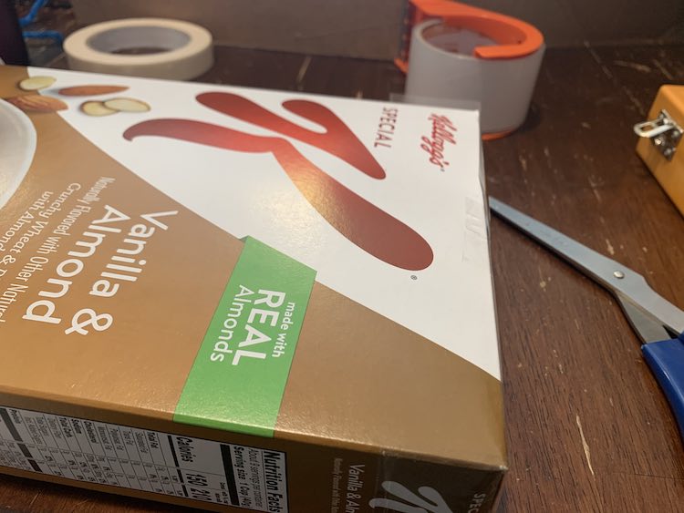 Image shows a closeup of the Kellogg's Special K cereal box with tape attached to the right side and sticking up from the back corner. Around the box sits blue-handled scissors, packing tape, masking tape, and a portion of the X-Acto knives box pokes in from the right. 