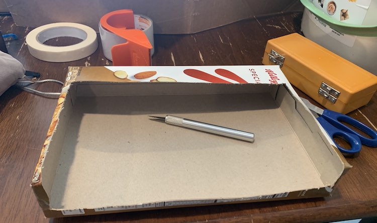 Image shows the box on it's side with most of the top and front cut away. There's a large stripe of box in the top back, a smaller section at the bottom front, and a smaller lip along the sides. The silver X-Acto knife sits in the opened box and the rest of the tools sit scattered around the box. 