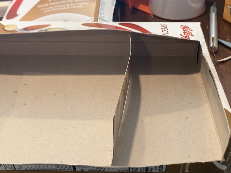 Image shows the cereal box on it's side with the top and front cut off. Inside a strip is loosely placed vertically to act as a divider making one large and one small space in the box. 