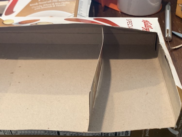 Image shows the cereal box on it's side with the top and front cut off. Inside a strip is loosely placed vertically to act as a divider making one large and one small space in the box.