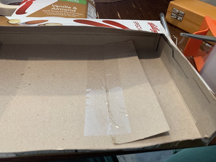 Image shows a closeup of the divider within the opened cardboard box. It's laying flat so it blends in with the box and there's a clear shiny piece of tape along it's left side taping it to the box. 