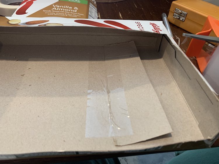 Image shows a closeup of the divider within the opened cardboard box. It's laying flat so it blends in with the box and there's a clear shiny piece of tape along it's left side taping it to the box.