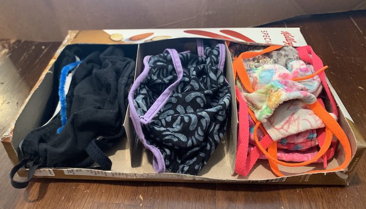 Image shows the just made organizer with all the masks placed carefully in piles inside. The far left shows a pile of black masks with a blue pipecleaner on top available to be inserted into a mask. The center shows my masks with a black and marble decorated one on top with lavender bias tape. The far right shows a pile with brighter colors and patterns along with neon shoelaces as a tie. 