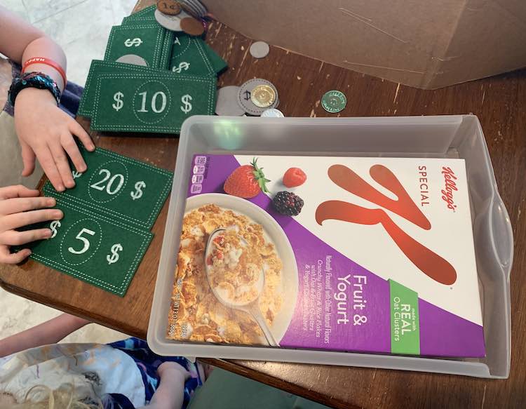 Image shows the plastic drawer sitting on the table with the Kellogg's Special K cereal box laying inside. Off to the side Ada and Zoey are standing by the table organizing the bills and coins. 