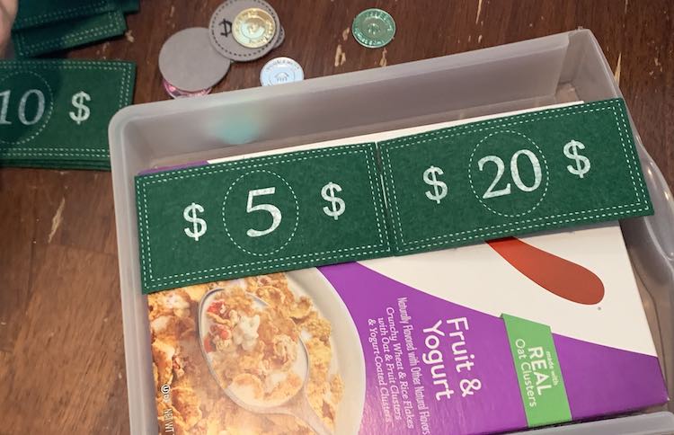 Image shows the cereal box within the plastic drawer again. There's a $5 and $20 bill side by side. The $5 is against the back of the drawer while the $20 bill beside it overlaps the side of the box and lays over the top of the drawer's front. 