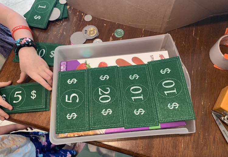 Image is take from above looking down at the plastic drawer. There are four bills laid side by side (5, 20, 10, and 10) going from the left to the right and slightly overlapping the front edge of the drawer. Off to the side Ada is organizing a pile of $5 bills while Zoey is seen, slightly, beside her.