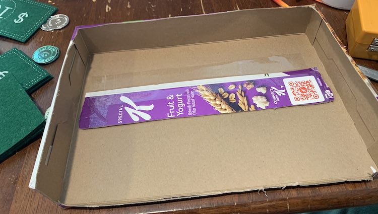 Image shows the opened cardboard box set on a table with a strip of cereal box placed printed side up across most of the width of the box. It's taped down along the top side.