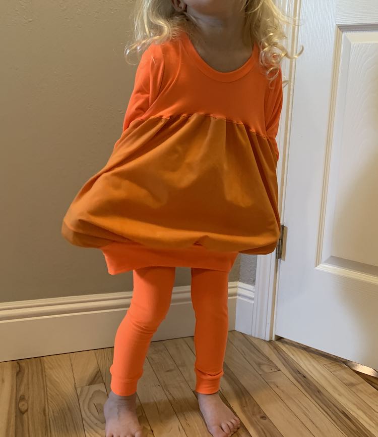 Front view of the pumpkin top with the sides spread out with Zoey's hands hidden in the pockets. The leggings are worn underneath the top.