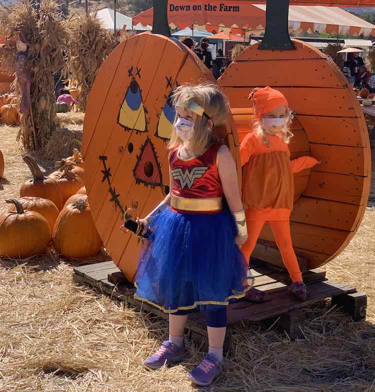 Both girls standing in front of another wooden pumpkin-like display with pumpkins laid out behind it. Ada stands in front looking off to the side while Zoey stands behind looking in the other direction.