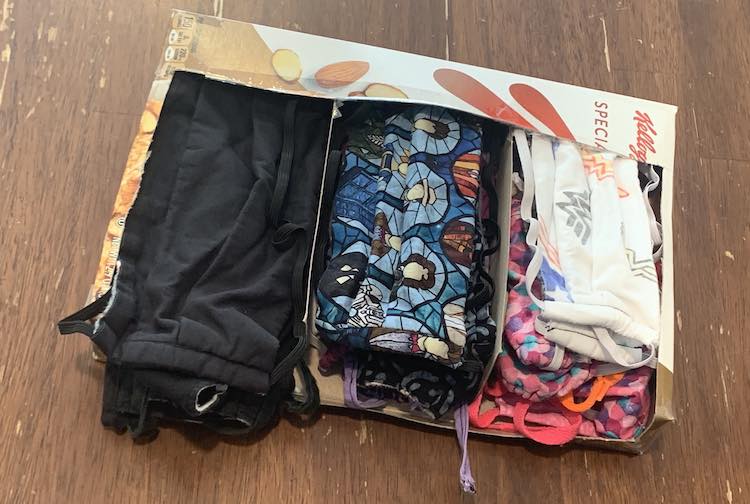 Image shows a cereal box with the top and front count out with two dividers so there's three separate sections for mask. In the left is a pile of black masks, while mine are in the center, and the kids' masks are on the right.