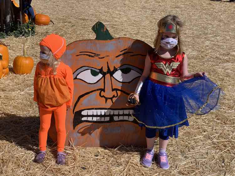 Image shows a painted wooden pumpkin with a Zoey costumed pumpkin and an Ada Wonder Woman standing by it. Around it lays straw on the floor and actual pumpkins off to the side. 