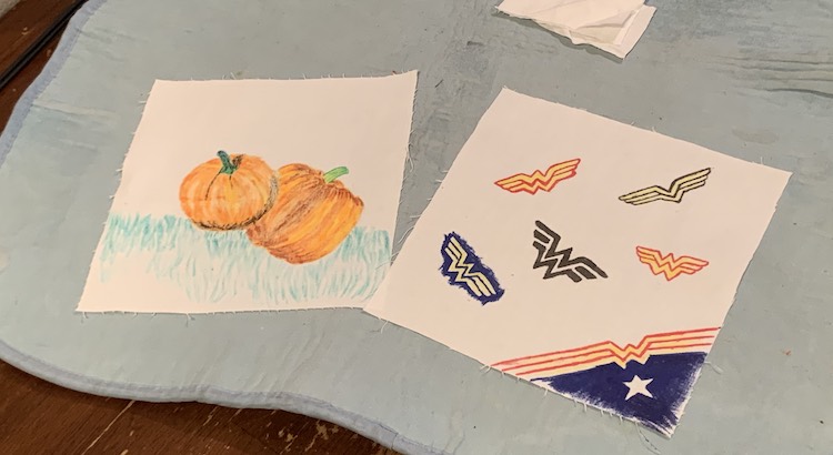 Image shows a blue ironing mat on the kitchen table with two rectangles laid out. In the background are a couple scraps of fabric, stacked, for the sides of the mask. The left rectangle shows two pumpkins with some grass below while the right one shows six Wonder Woman logos I copied from my internet search results.