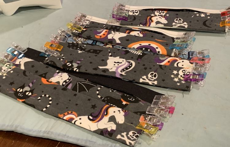 All four masks are laid out on the blue ironing mat on the table. The top of each shows a small strip of the outer fabric as it was folded in. Other than that they all have the same grey starred fabric with horned unicorns, pumpkins, cats, and skulls tossed around. The pleats make it appear like those old paper images that you fold in to change the image.