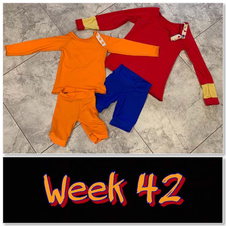 Image shows the finished swim garments laid out on the kitchen floor with a black rectangle below showing the words "week 42" in orange and red. The swim garments are composed of an orange rash guard,  orange shorts, red rash guard with yellow bands, and blue shirts. 