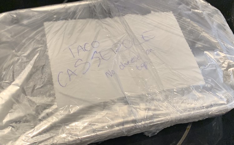 Image is taken from above showing a casserole dish covered in tinfoil, a piece of napkin saying "taco casserole no cheese on top". and plastic wrap. It sits on the top of the stove. 