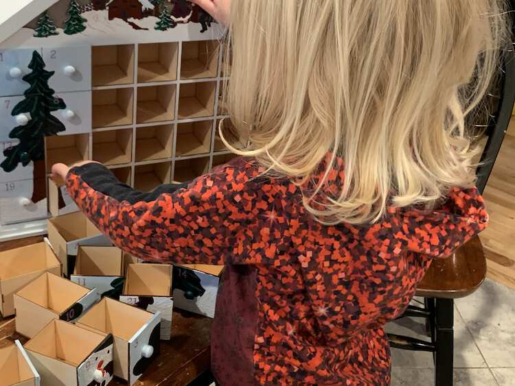 Image shows the back of Zoey in a Christmas red shirt as she builds the advent calendar puzzle. The calendar shows most of the left tree and the rest of the painted drawers sit on the table. 