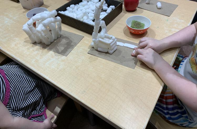 Image shows a table that two kids are sitting at making a biodegradable packing peanut sculpture on a piece of cardboard. In the middle sits a tray of more packing peanuts and beside them sits a bowl with a damp sponge inside. 