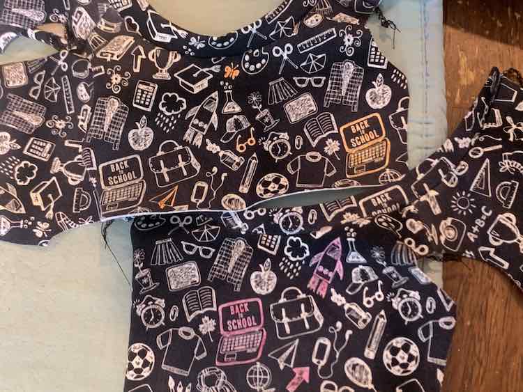 Image shows two swatches of black fabric with white school-related doodles on it. There's a few seams but they're flattened over the table and an ironing mat. The top swatch has some of the doodles colored in with orange while the bottom one has three doodles colored in with pink.