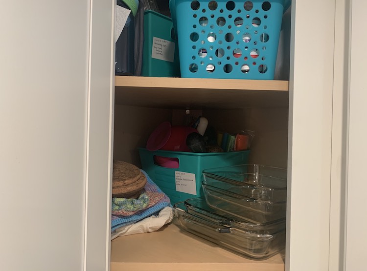 Image shows my opened corner cupboard. Inside you can see two shelves with the one bin, hidden behind, at the top and the other bin facing the other way in the back corner on the bottom shelf. Those two bins have the white sticker stuck under the handle. 
