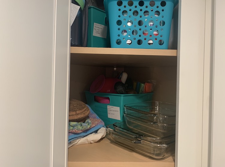 Image shows my opened corner cupboard. Inside you can see two shelves with the one bin, hidden behind, at the top and the other bin facing the other way in the back corner on the bottom shelf. Those two bins have the white sticker stuck under the handle.