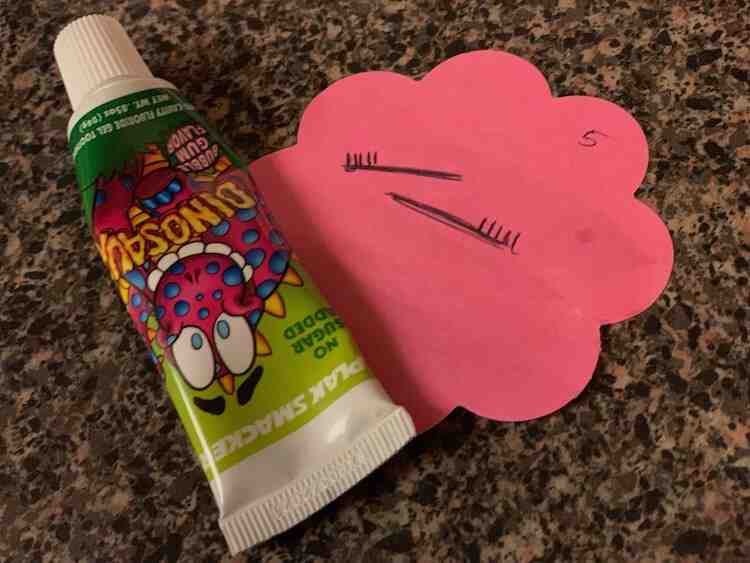 Image is showing a small section of the counter with a pink post-it not with two drawn toothbrushes. Slightly overlapping is a small tube of toothpaste with a dinosaur on it. 
