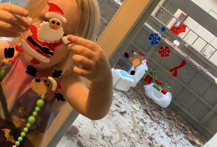 Image shows a window gel cling Santa being held up in front of Ada's face. Behind her is the patio door with some of the other window gel clings already attached on. 