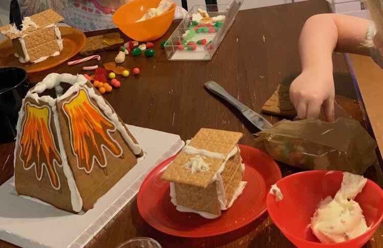 Image shows a table with a gingerbread volcano spouting colorful sugar paper lava. Behind and to the side of it the kids are seated with a graham cookie and icing house and candy to decorate it with.