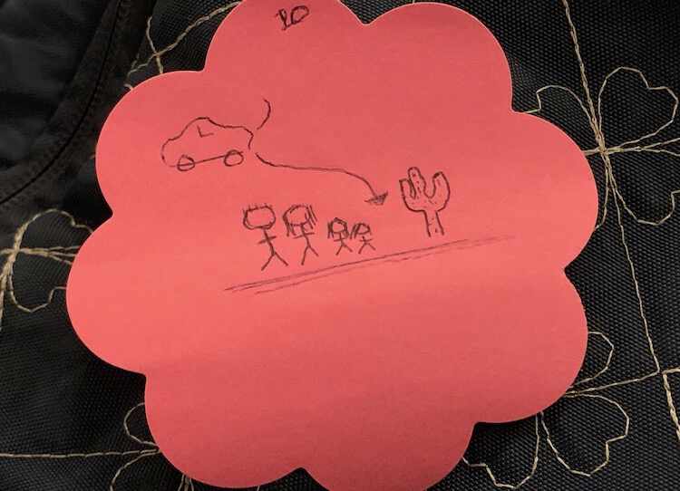 Image shows a pink flower post-it note on top of my diaper backpack. On it is drawn an arrow with a car mid-way to the destination at the tip of the arrow. On it is a cactus, the ground, and four figures of proper heights. 