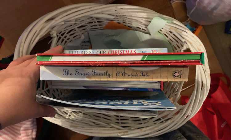 Image take from above looking down at a white wicker basket filled with Christmas themed books sitting upright inside it. 