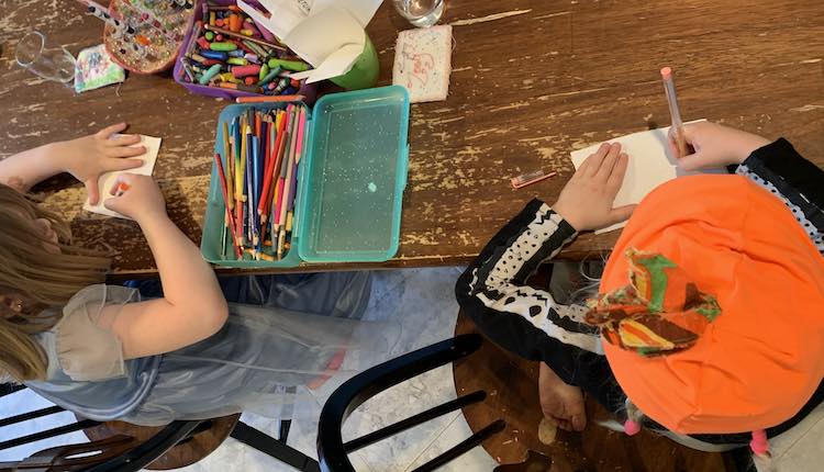 Image shows the kids sitting beside each other at the kitchen table. Between them sits a container of crayons, a container of pencil crayons, a marker holder, and a cup of blank stickers. 