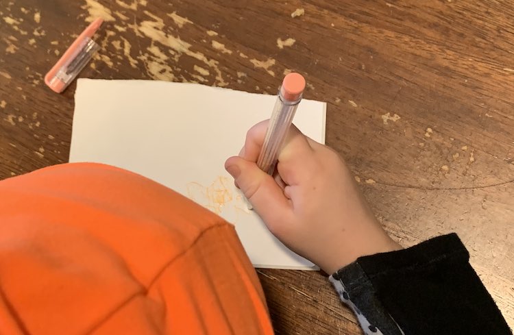 Image is take from above Zoey looking down at the table. You can see part of her pumpkin hat and her hand while she doodles on the sticker with an orange-y peach gel pen. 