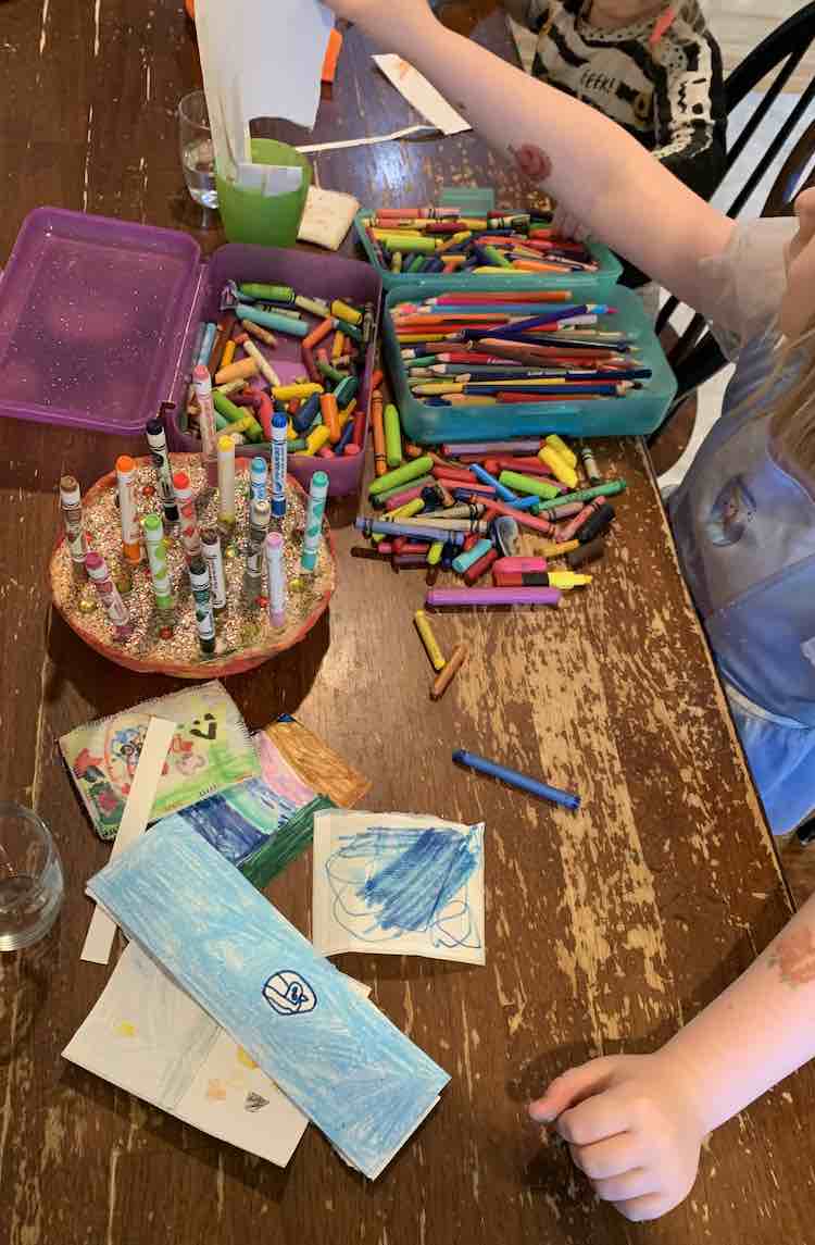 Image shows the table, stretching from the bottom of the image to the top. Ada is grabbing a new blank sticker to design another to add to her finished pile in front of her. Between the kids sits their shared art supplies. 