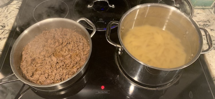 Image shows my stove with the front two larger burners being used and two glass lids behind them. The left saute pan has browned ground beef in it while the rightmost pot has just added penne pasta so the water hasn't yet come back up to a boil. 