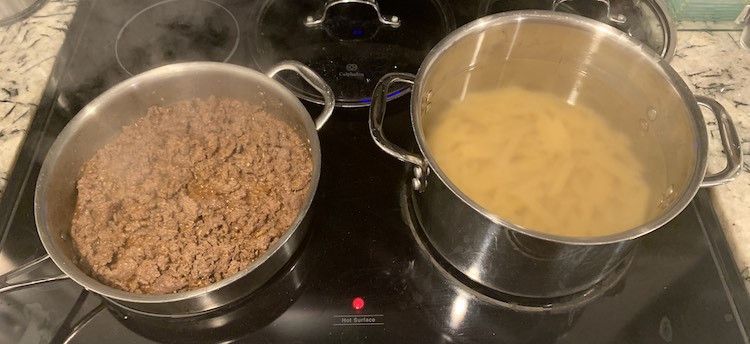Image shows my stove with the front two larger burners being used and two glass lids behind them. The left saute pan has browned ground beef in it while the rightmost pot has just added penne pasta so the water hasn't yet come back up to a boil.