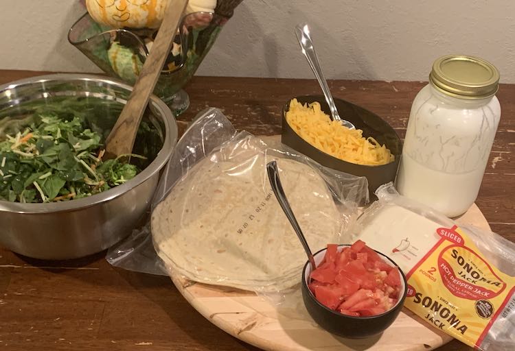 Image shows a wooden lazy Susan laden with a package of flour tortillas, a container of grated orange cheddar cheese, a glass mason jar a third way filled with white yogurt, a package of sliced hot pepper jack cheese, and a small black bowl filled with diced tomatoes. To the left sits a large metal bowl filled with greens and carrot pieces with salad spoons. In the back sits a glass bowl filled with decorated pumpkins. 