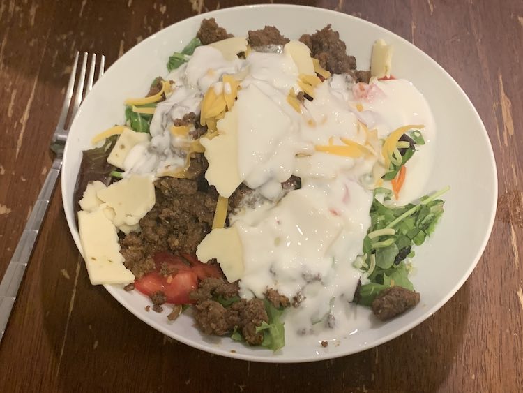 Image shows a white bowl with a fork laid out beside it. The bowl is filled with a variety of colors including green, brown, red, orange, and a lot white.