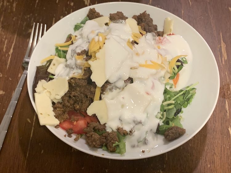 Image shows a white bowl with a fork laid out beside it. The bowl is filled with a variety of colors including green, brown, red, orange, and a lot white. 