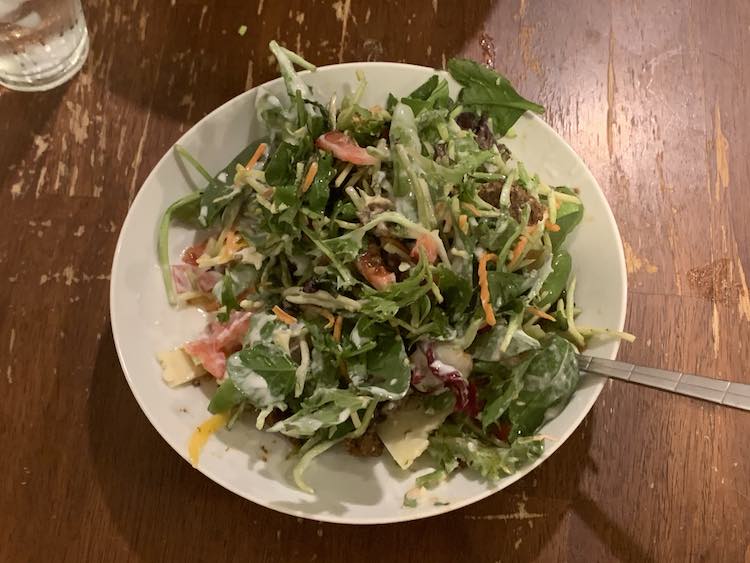 Image now shows a bowl filled with a more colorful looking salad and a fork sticking out of it with a water glass off to the upper corner. The bowl shows julienned carrots and cabbage, a salad mix, cheese, tomatoes, and yogurt 'dressing'.