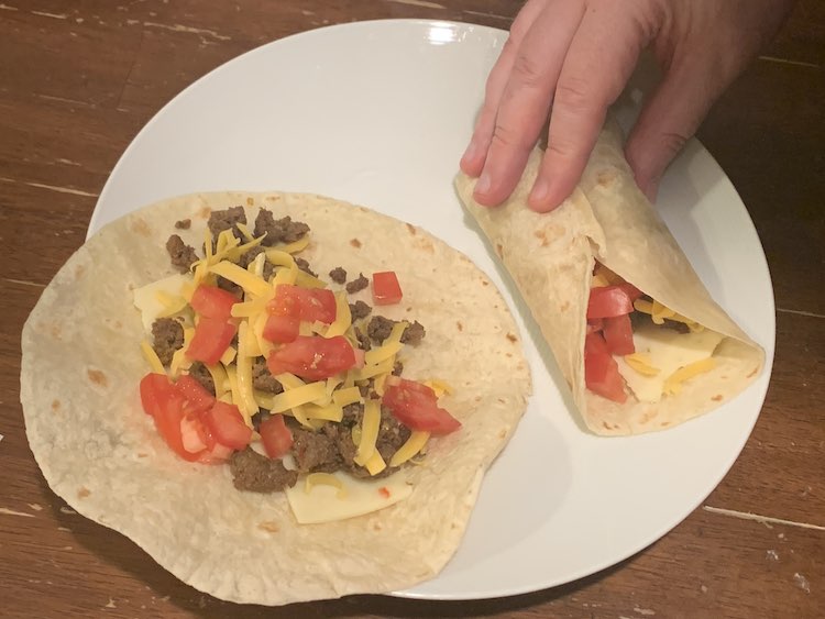 Image is focused on a white plate with an open wrap on the left and a hand holding the wrap on the right closed. Both wraps are filled with pepper jack cheese, taco meat, grated cheddar cheese, and diced tomatoes. 