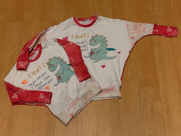 Image shows two long sleeved shirts laid out on a floor. The bottom band at part of the front of the sleeve has pink "Love" and hearts on it. The back of the sleeves, neckband, and cuffs are dark pink with hearts. The panel features a dragon, words saying "rawr! that means I love you in dinosaur", and several hand drawn shaded hearts in orange (left shirt) and red (right shirt). 