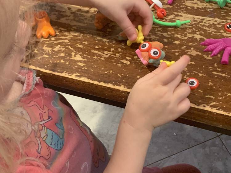 Image shows Zoey holding the hands of a little orange and blue monster with giant looking eyes, pink feet, and yellow hands.