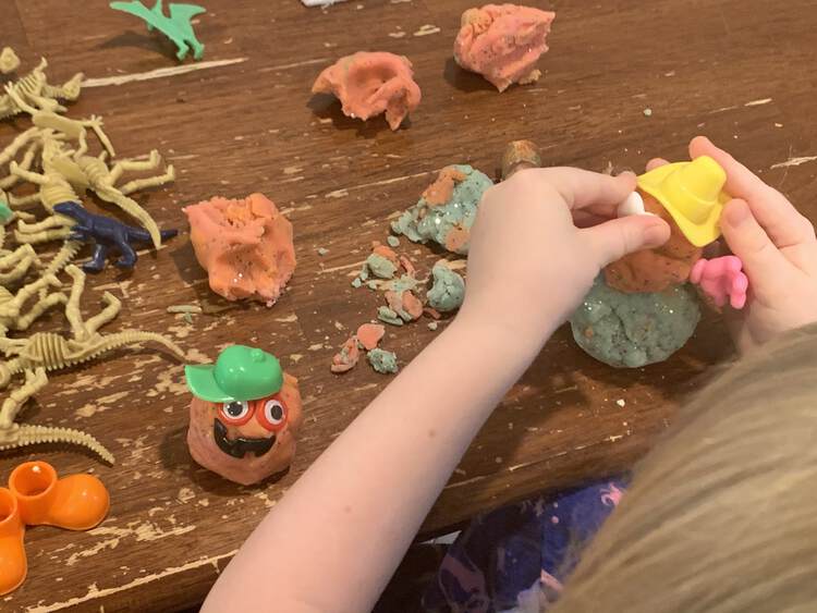Image shows Ada adding eyes to her cowboy hat wearing playdough snowman. A little circled monster sits beside and an assortment of plastic dinosaurs and playdough pieces are scattered behind.