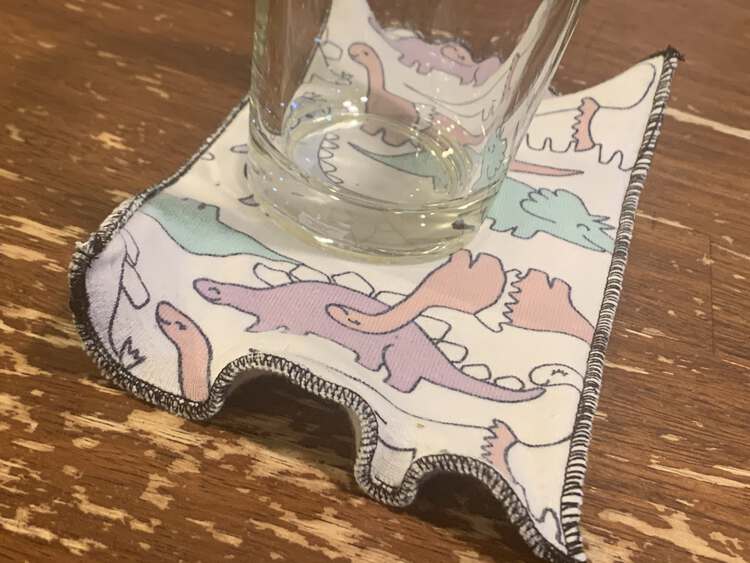 Image shows an empty glass sitting on a serged fabric swatch on the kitchen table. The edge is stitched in black to go with the black outlined dinosaurs on the fabric. The edge closest to you is folded but the sides are flat. 